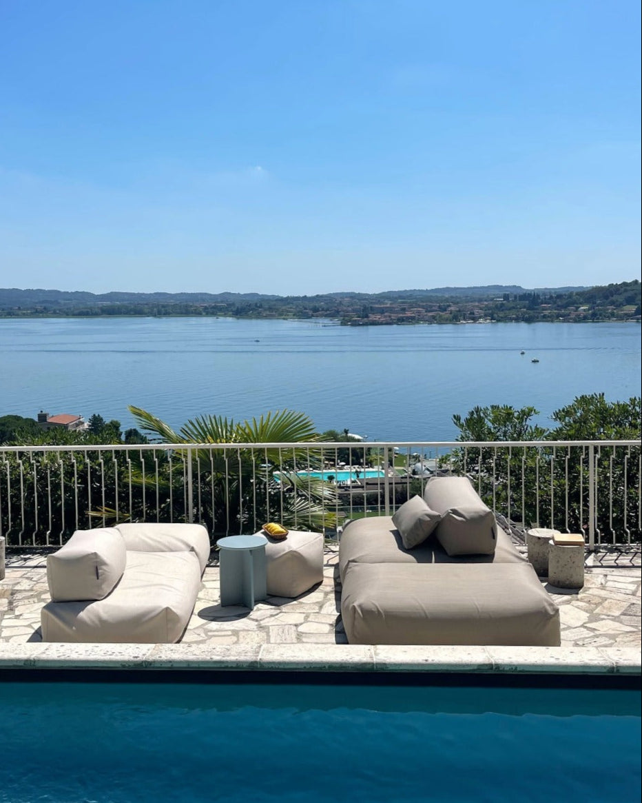 An einem Pool stehen zwei Poolsofas (ein Double in Biege, ein Single in Perlmutt) gegenüber als Outdoorlounge und ein Pouf in Small und wird als Tisch genutzt. $ Poolsofa Lounge | Perlmutt & Beige