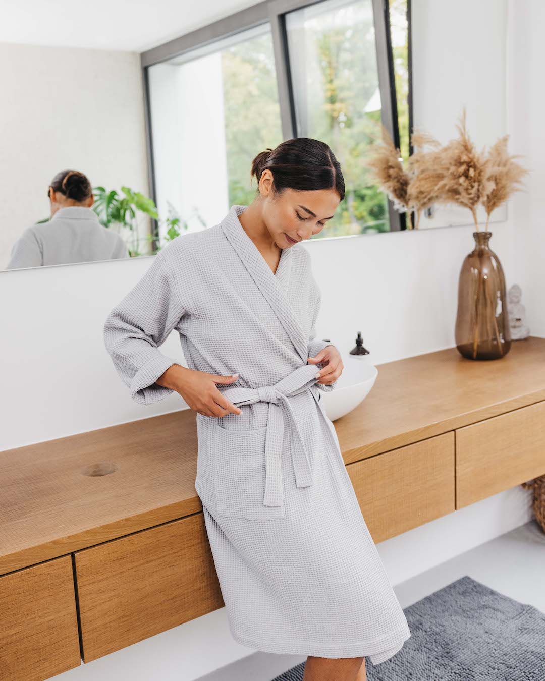 Frau steht im Bademantel (Farbe Cloud Hellgrau)) angelehnt ans Waschbecken und bindet die Schleife ihres Bademantels.