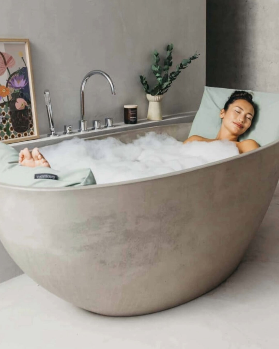 Eine Frau liegt entspannt in einer freistehenden Badewanne, sie lehnt sich an ein Badesofa Rückenkissen Large in der Farbe Grün, ihre Füsse ruhen auf einem Fusskissen, ebenfalls in Grün. 