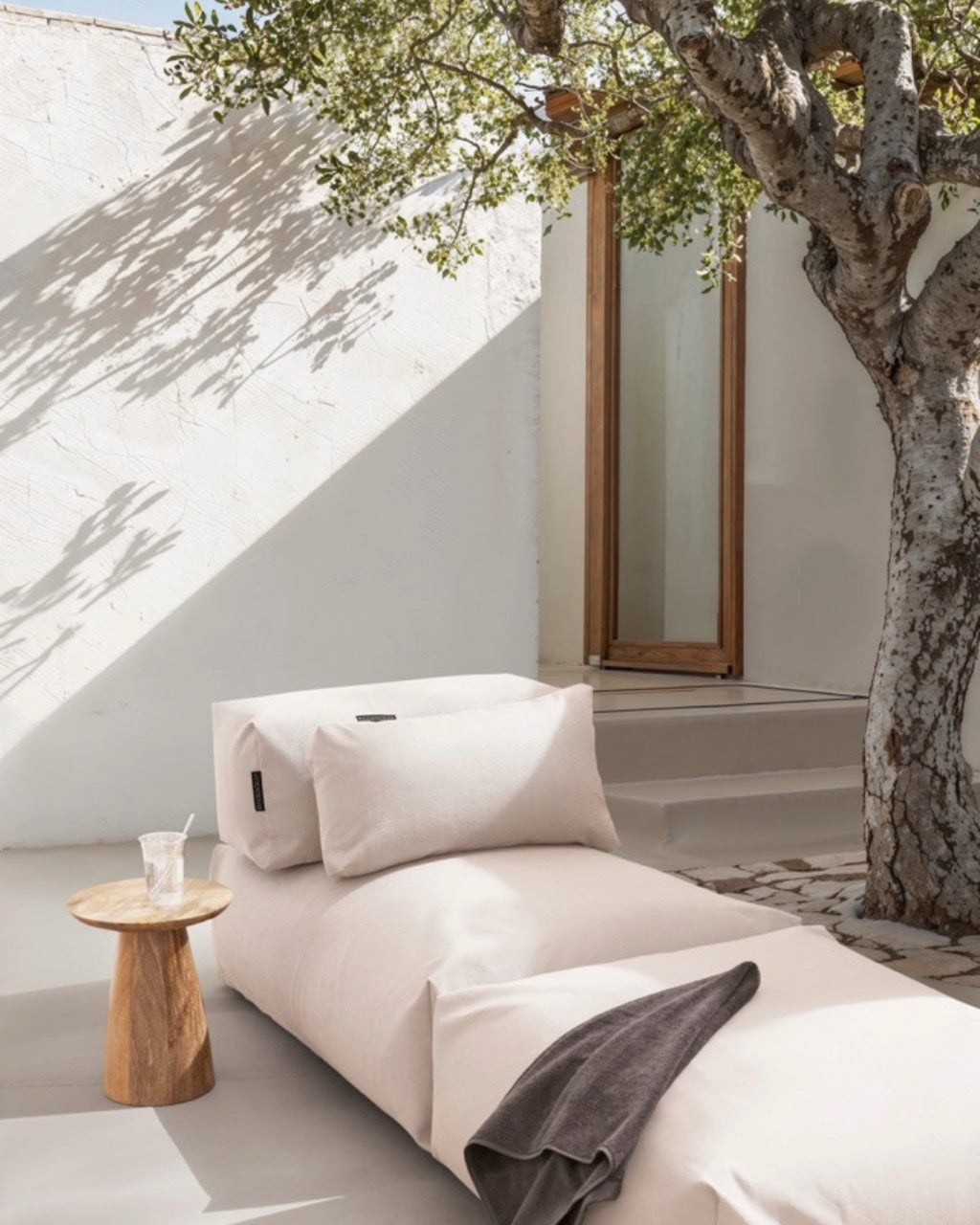 Unter einem Baum steht ein Poolsofa Singel in Perlmutt, daneben ein kleiner Holztisch. Das Rückenkissen und Relaxkissen sind auf dem Poolsofa drapiert. #farbe_perlmutt $ Poolsofa Single | Perlmutt